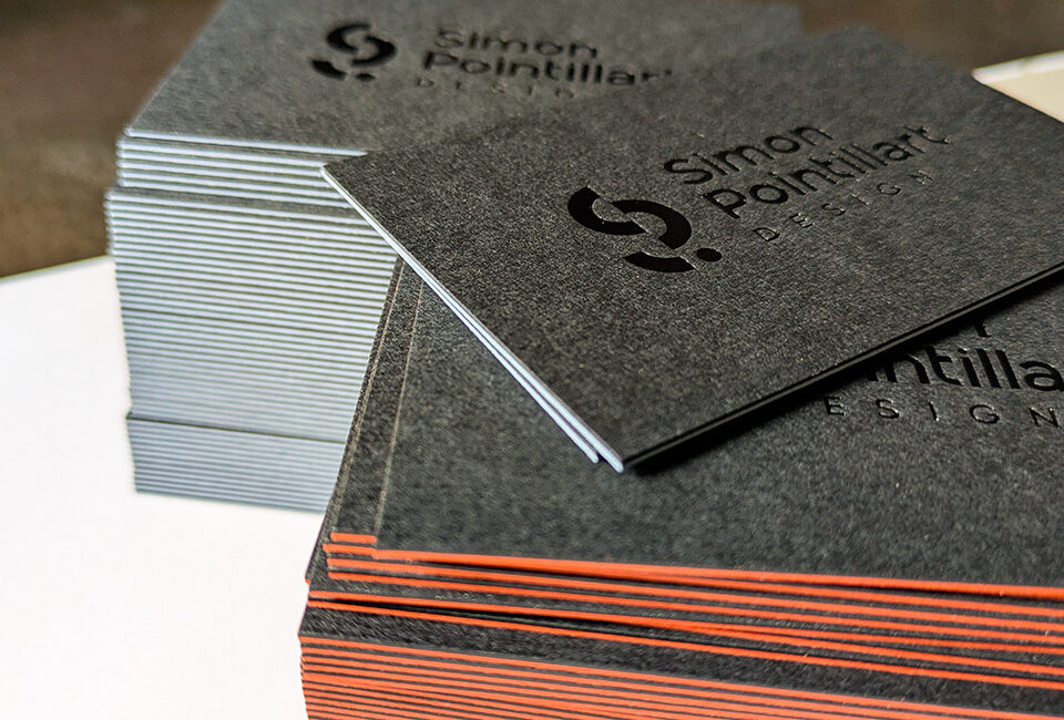 Cartes de visite haut de gamme pour architecte d’intérieur, avec marquage à chaud noir brillant sur papier anthracite mat et tranches orange et bleu clair.